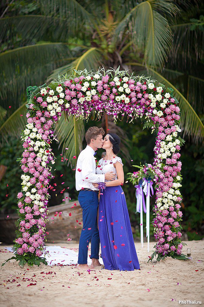 Возле свадебной арки, украшенной белыми и сиреневыми цветами, целуются молодожены, жених в синих брюках и белой рубашке держит за - фото 589754 PhoThai - свадьбы и фотосессии в Таиланде
