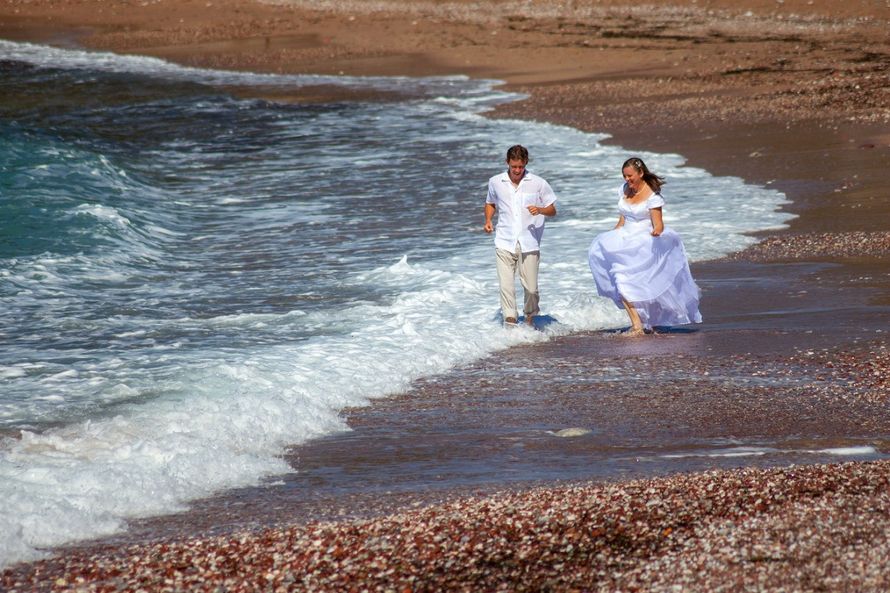 По пляжу, идут жених с невестой, она в белом, длинном платье, держит юбку руками, он в белой рубашке и серых брюках - фото 1579895 Свадебный фотограф Arina Barina