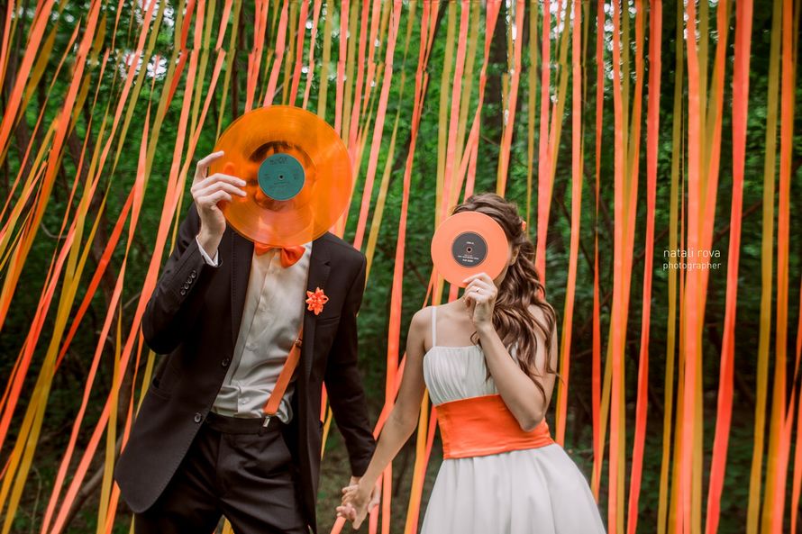 На фоне оранжевых лент, стоят жених и невеста, прикрыв лица оранжевыми бумажными цветами - фото 2625293 Фотограф Натали Рова