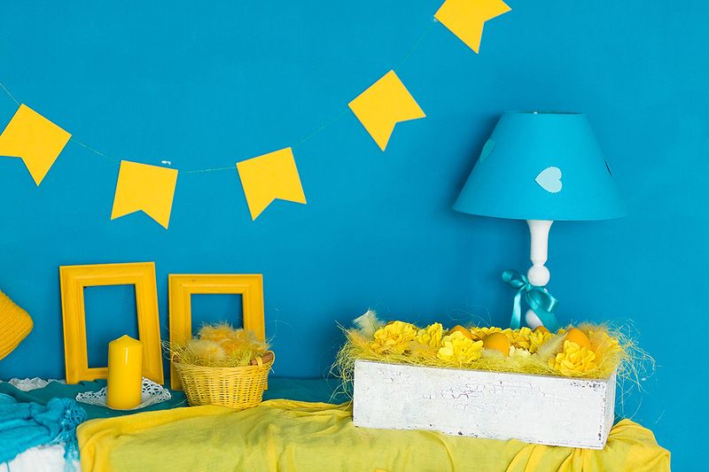 Возле голубой стены, украшенной жёлтыми флажками стоят голубая лампа, жёлтые рамки и корзинки - фото 1231373 Pro-decor - студия оформления