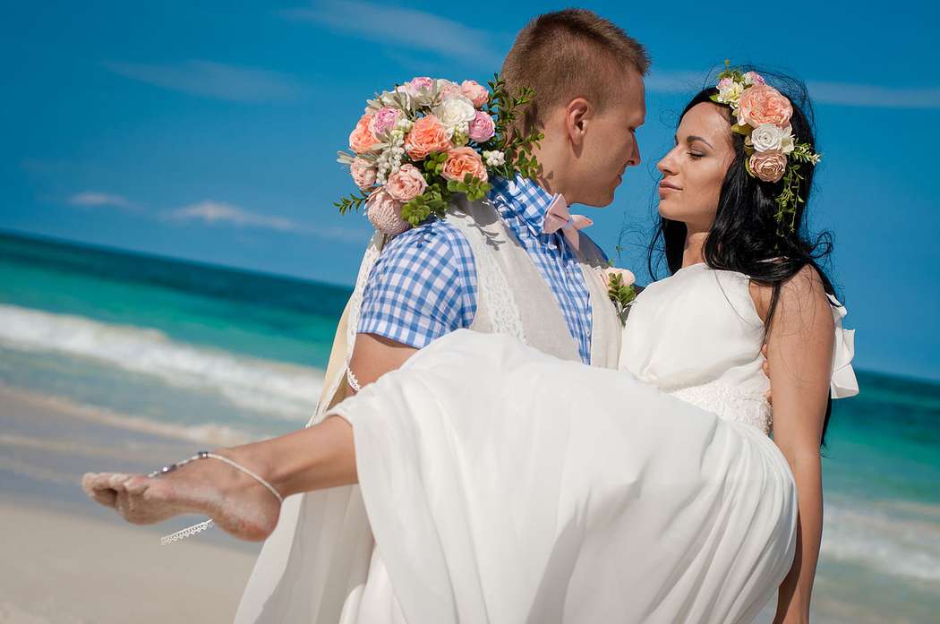 На тропическом побережье стоят влюбленные, жених в светлых бриджах, жилетке и клетчатой рубашке держит на руках невесту в белом - фото 2703956 Danaysha