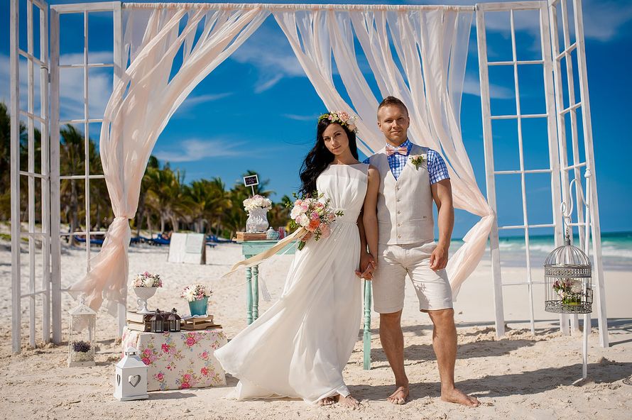 На пляже на фоне свадебной арки, оформленной в виде огромного окна, стоят молодожены, взявшись за руки, невеста в белом длинном - фото 2703960 Danaysha