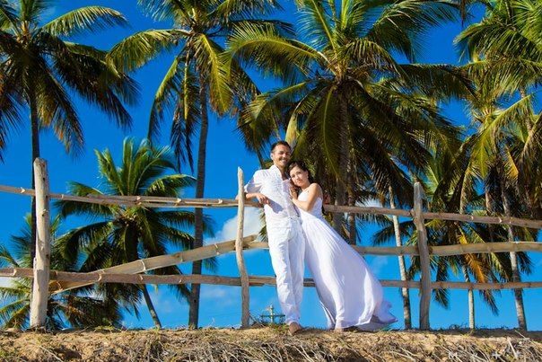 На фоне пальм, возле ограждения, стоят молодожены, невеста прижалась к жениху в белом, длинном, платье, он в белом костюме - фото 1859907 Свадебный фотограф Макс Клевер