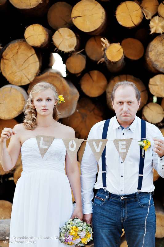 На фоне брусьев дерева, держа в руках табличку-надпись, стоят жених и невеста - фото 2739005 Свадебные фотографы StoryOfMoments 