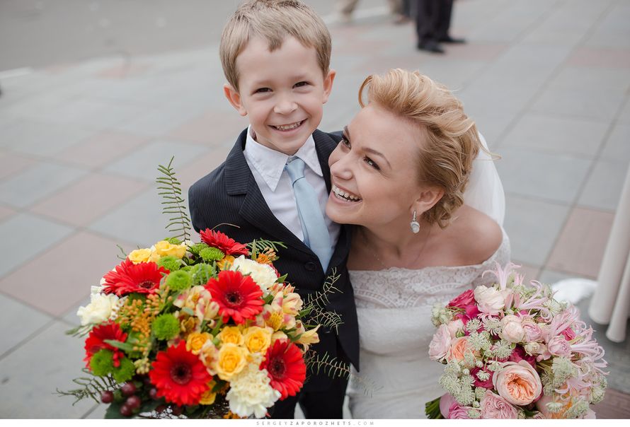 Мальчик, улыбаясь обнимает невесту  в в костюме с белой рубашкой и голубым галстуком и держит букет из красных гербер - фото 2358418 Анна Карташова