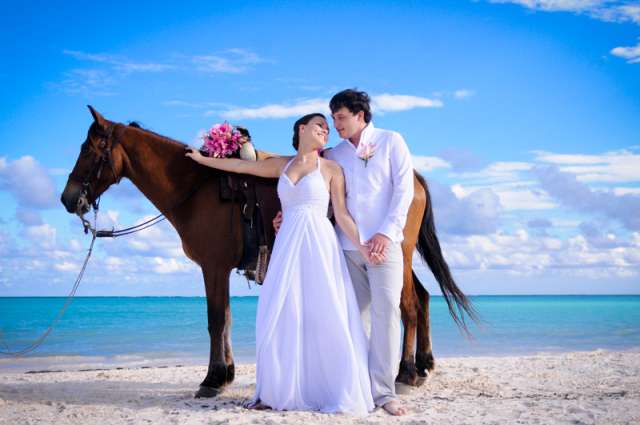 На побережье возле лошади стоят молодожены, жених в белых брюках и рубашке держит за руку невесту в белом платье, она повернула - фото 2292292 Туристическое агентство Англетер