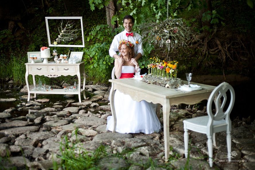 На фоне зеленых кустов, возле белой тумбочки, за белым столом сидит невеста, за ней, обнимая ее за плечи, стоит жених - фото 2581185 Декоратор Мария Демьянова