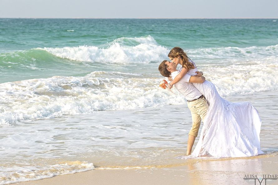 На песчаном побережье жених в светлых брюках и белой рубашке держит за талию невесту в белом платье, слегка приподняв, в руке у - фото 2766645 "Tytprod" - фото и видеосъёмка 