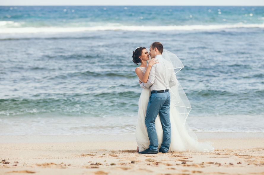На пляже стоят жених и невеста, они обнимают друг друга, на ней длинная прозрачная фата и белое длинное платье, он в белой - фото 3517821 Happiness :)