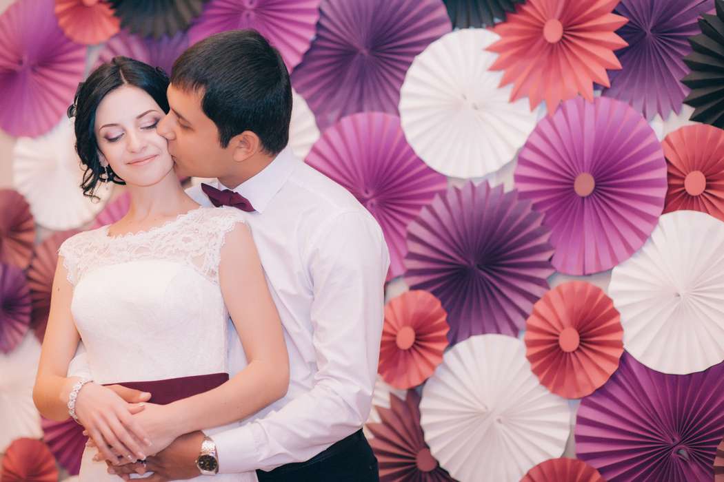 Жених целует невесту на фоне бумажных бело-розово-фиолетовых зонтиков - фото 3331749 Свадебное агентство "Lucky Wedding"