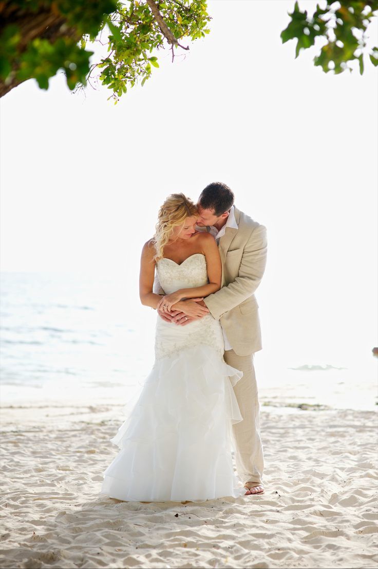 На пляже на фоне океана жених в светлом костюме обнимает за талию и целует невесту в открытом белом платье "русалка" - фото 3380215 Holiday Vibes Photo фотосессии в Мексике