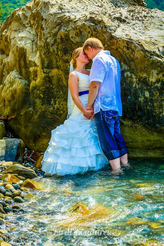 На пляже стоят рядом с камнем жених и невеста, взявшись за руки, он в голубой рубашке и синих брюках, она в открытом, пышном - фото 3438363 Свадебный фотограф Кирилл Бондарев