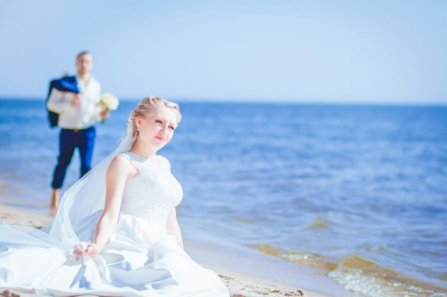 На пляже, невеста сидит на песке, в фате и белом платье,  жених идет в белой рубашке и синих брюках, с букетом цветов - фото 3555583 Фотограф Макс Мотайлов