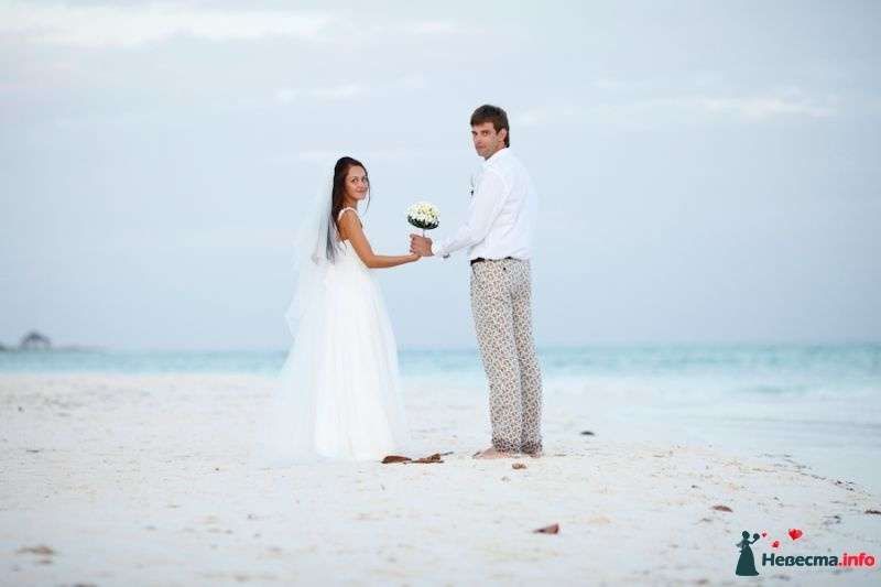 На песчаном пляже стоят молодожены, жених в светлых брюках и белой рубашке и невеста в белом платье с фатой держат в руках букет - фото 374242 Акварель