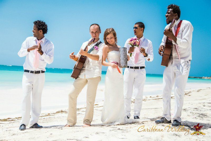 На пляже стоят гости с цветами и гитарой в белых костюмах, жених стоит в белой рубашке и брюках, играет на гитаре, невеста стоит - фото 2811207 Caribbean Wedding - свадьба в Доминикане