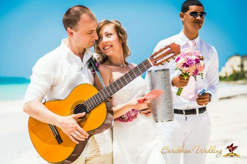 На пляже стоит гость с цветами  в белом костюме, жених стоит в белой рубашке и брюках, держит в руке гитару, невеста стоит в - фото 2811209 Caribbean Wedding - свадьба в Доминикане