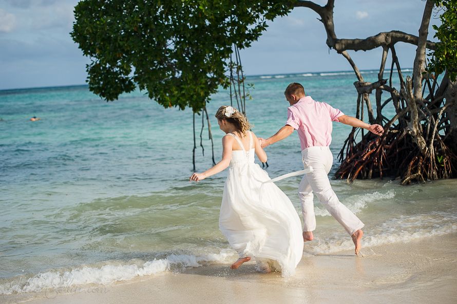 На тропическом пляже, взявшись за руки, забегают в воду молодожены, жених в белых брюках и розовой рубашке, невеста в белом платье - фото 566742 Valentinka~@~